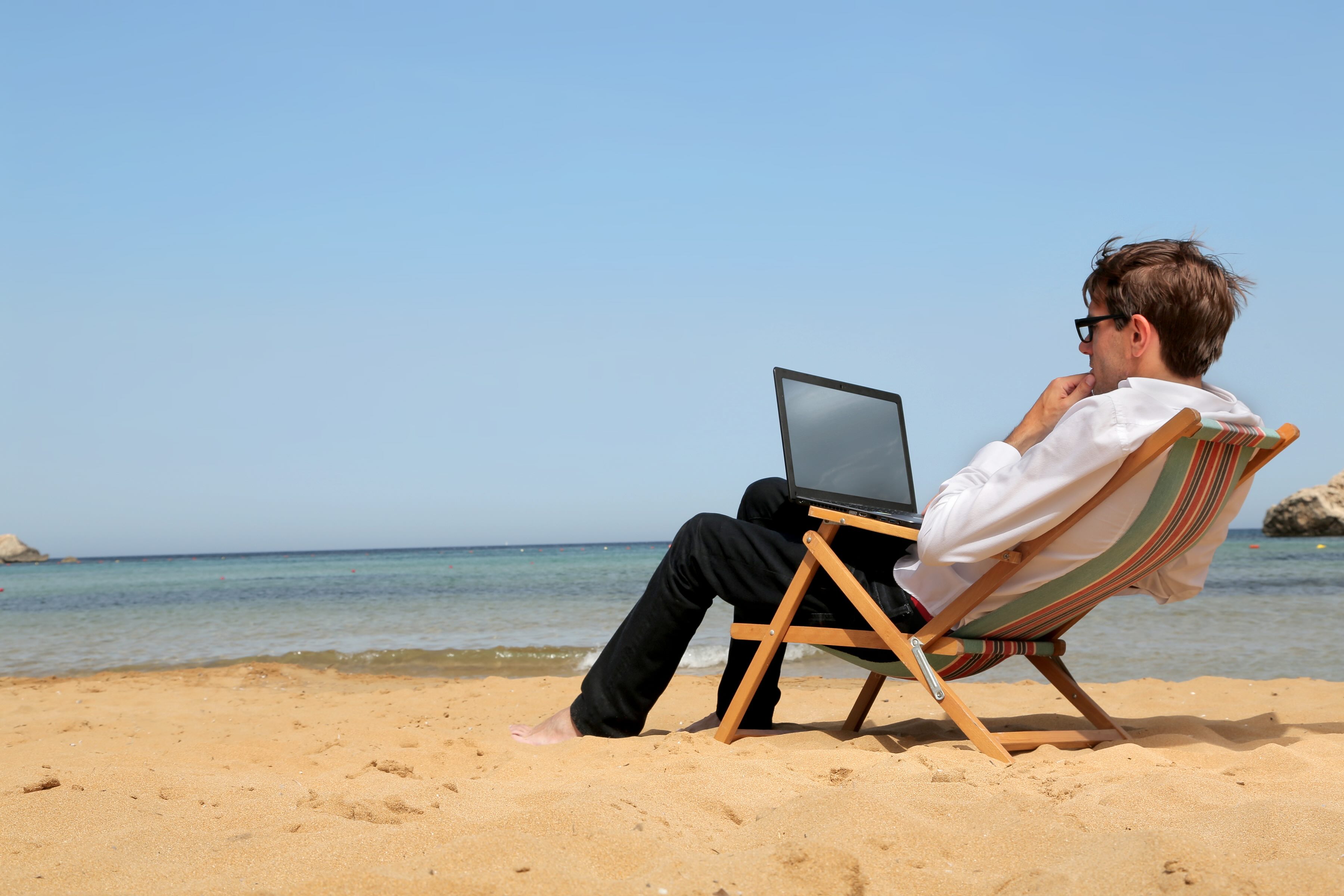 Man working on a laptop in a deckchair at the beach, indicating the flexibility with which exams may be delivered online